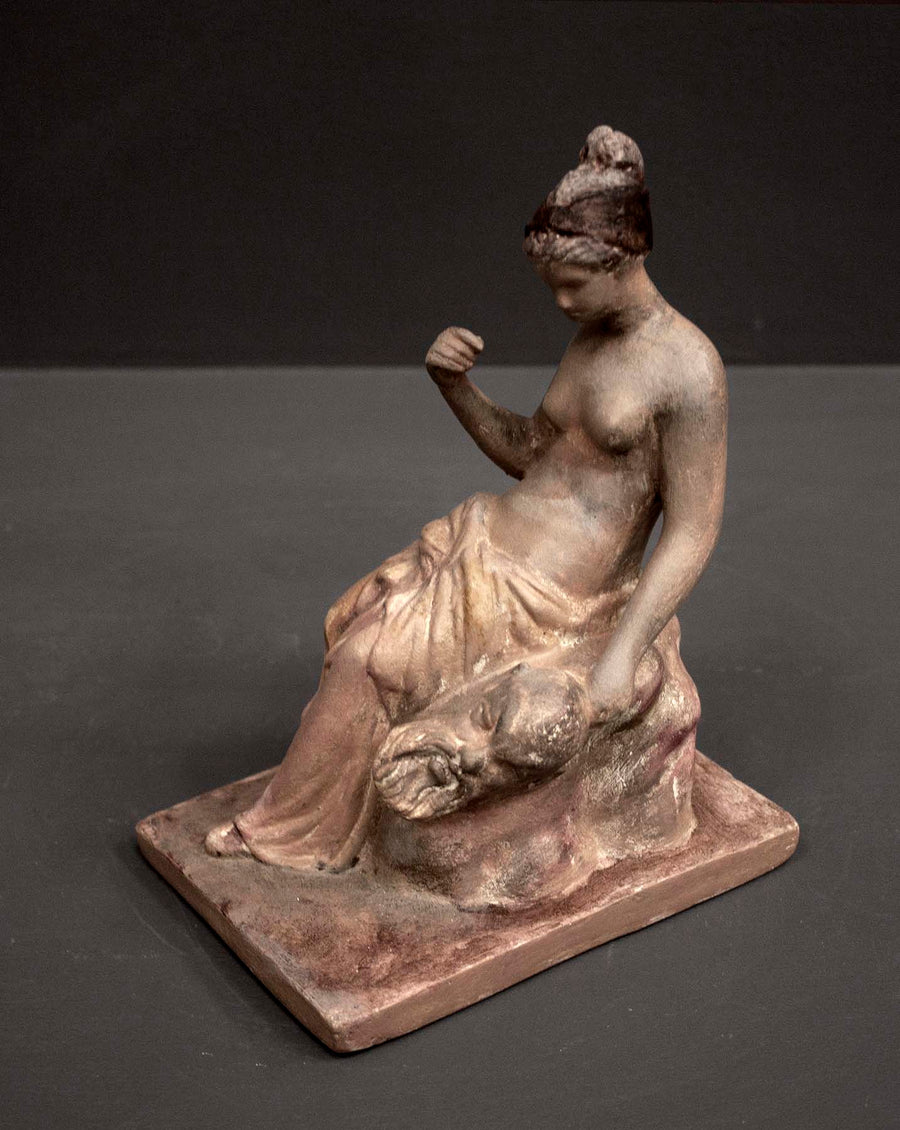 photo of plaster cast sculpture of female half nude with pink robes seated and holding a mask, set against gray background