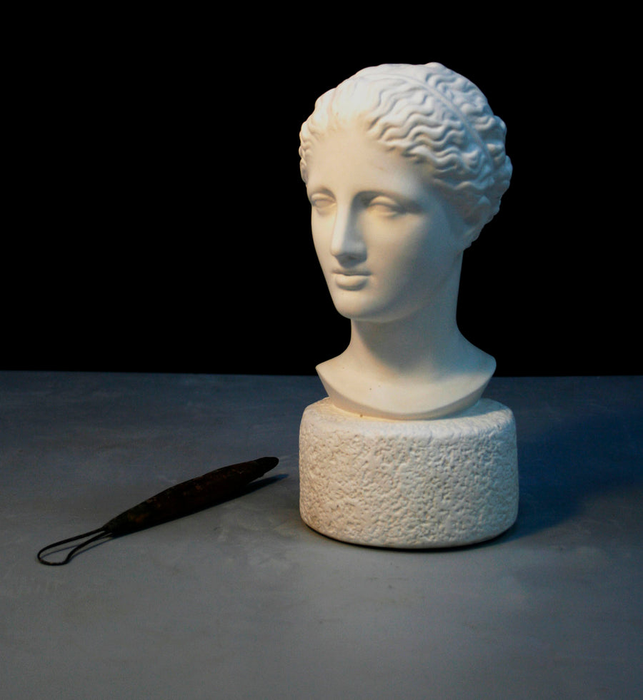 photo of plaster cast of ancient female bust, namely the goddess Diana, with a gray and black background and sculpting tool beside it