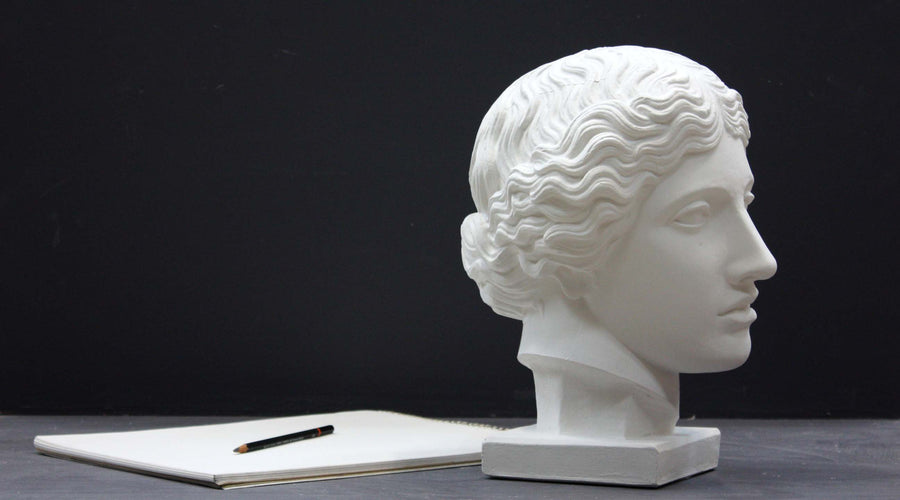 photo of plaster cast of sculpture of an Amazon head- female with pulled-back wavy hair on dark gray surface with sketchbook and pencil beside it