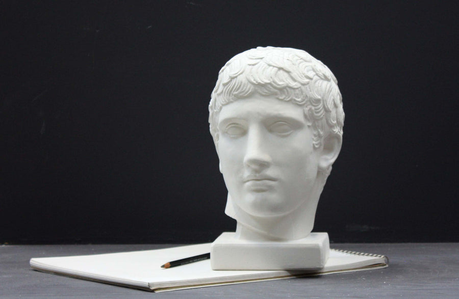 photo of plaster cast of sculpture of male head with flat, curly hair on top of sketchbook along with a pencil on dark gray surface