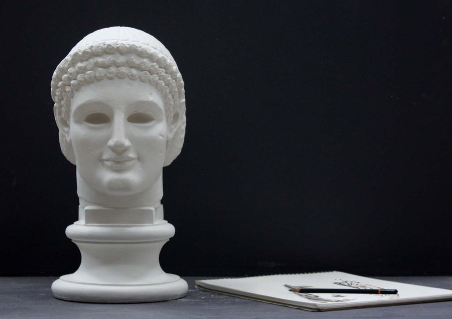 photo of plaster cast sculpture of archaic Greek male head with hair in fillet and ringlets on dark gray surface with sketchbook and pencil beside it