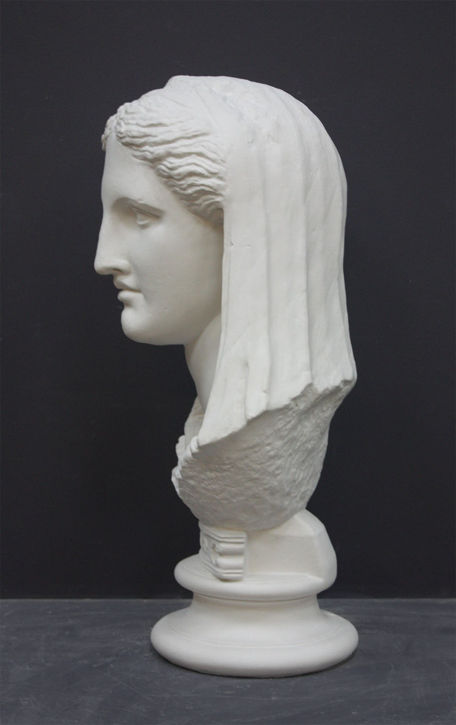 photo of white plaster cast of ancient sculpture of female head with curls and wrap, namely Demeter, on socle base on dark gray background