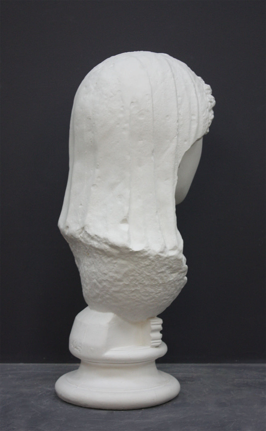 back view photo of white plaster cast of ancient sculpture of female head with curls and wrap, namely Demeter, on socle base on dark gray background