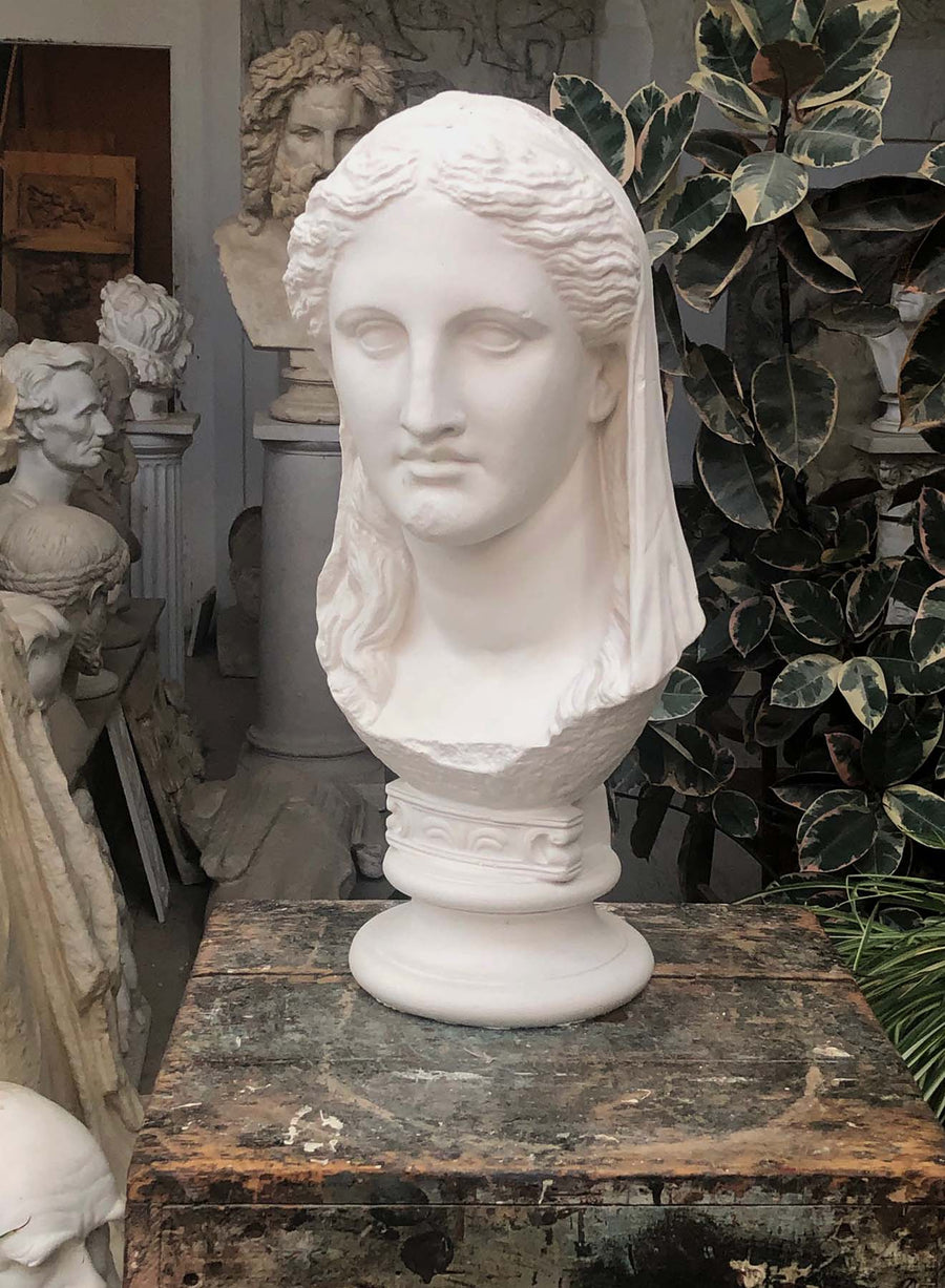photo of white plaster cast of ancient sculpture of female head with curls and wrap, namely Demeter, on socle base on wood table with greenery and sculptures behind