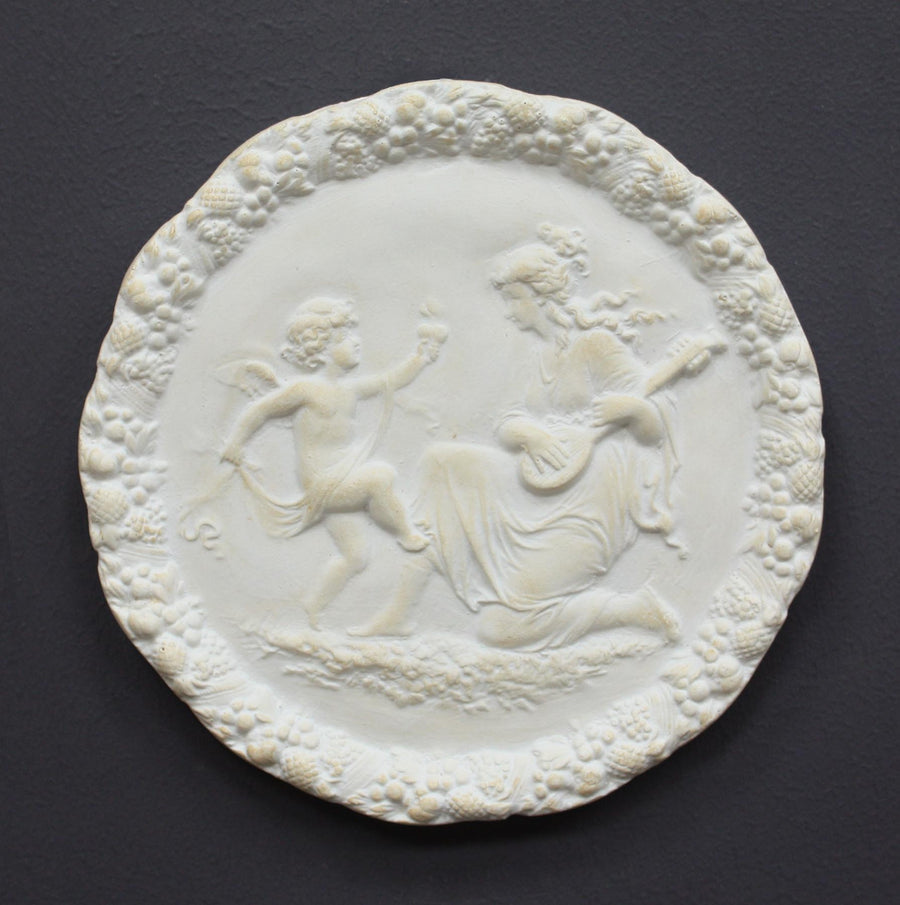 photo of white plaster cast relief sculpture of a child dancing and a female figure playing an instrument against dark gray background