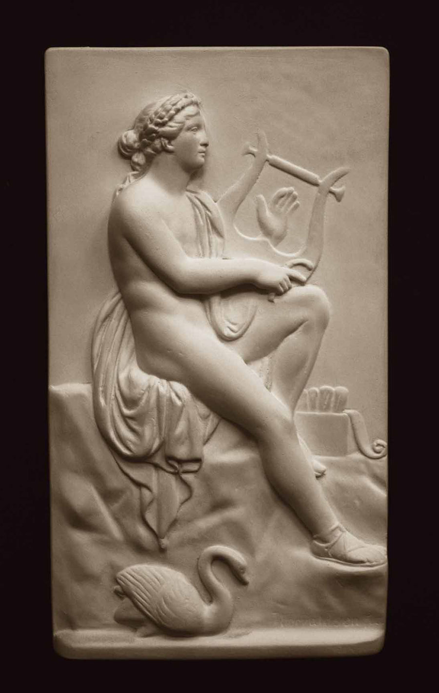 photo with black background of plaster cast sculpture relief of man, namely the god Apollo, seated on rock and playing a lyre