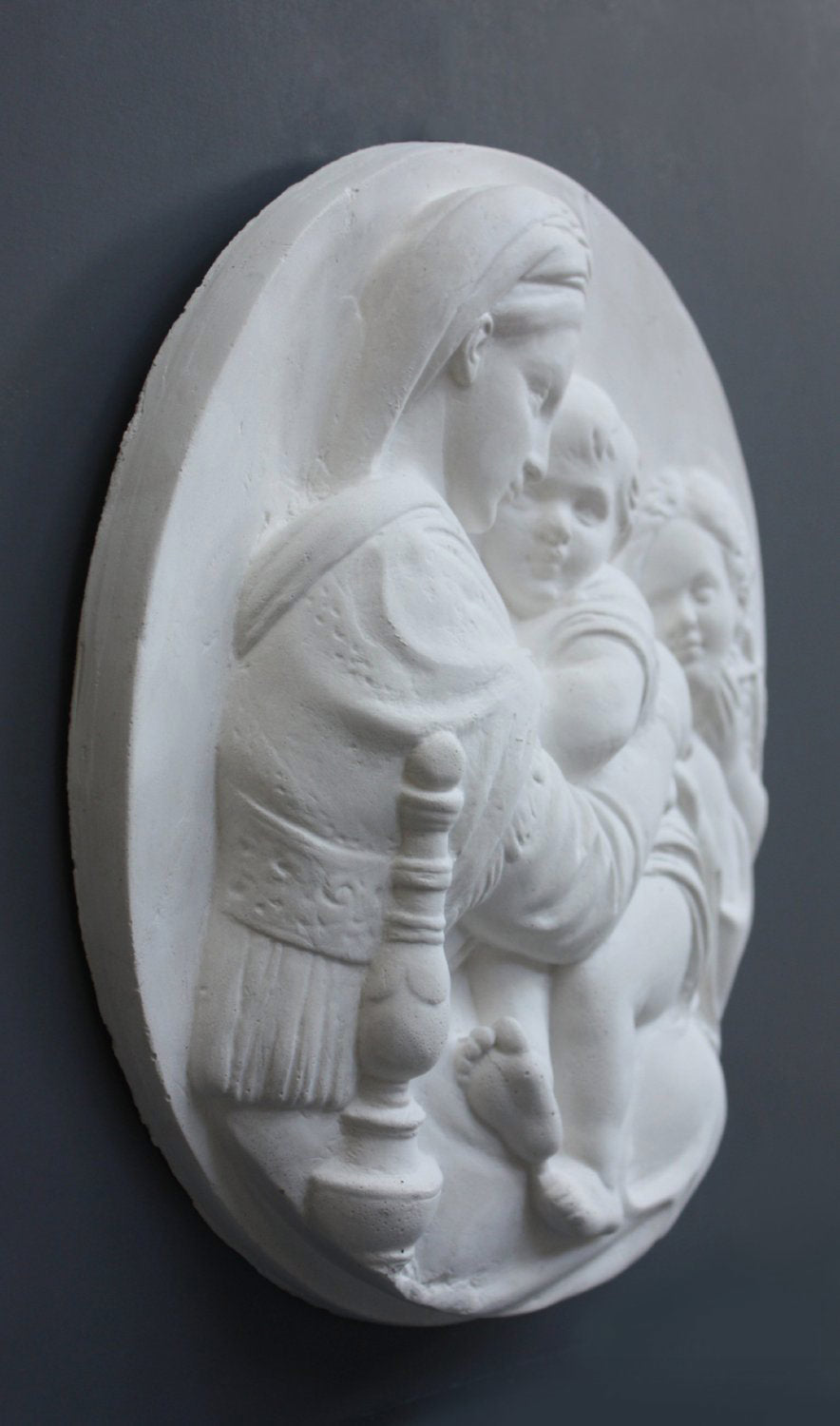 three-quarter photo of relief plaster cast featuring the Madonna, Child, and St. John and hanging on a dark-colored wall