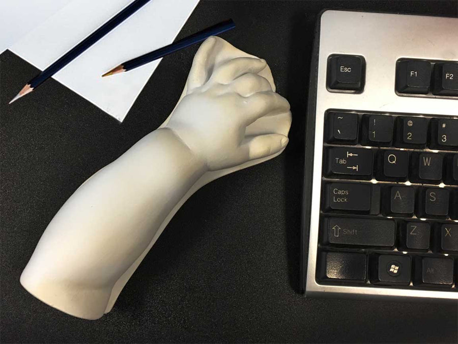 photo of white plaster cast sculpture of baby lower arm sitting on a desk between a keyboard on the right and a sheet of paper and two drawing pencils on the left