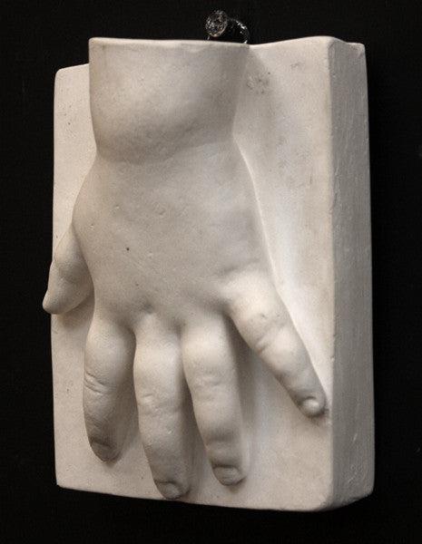 photo of off-white plaster cast sculpture of baby hand on panel against black background