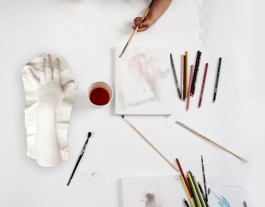 Photo of top of white table with child's hand holding a paintbrush, brishes and pencils and sketchpad scattered, with a plaster cast of a female hand to the left