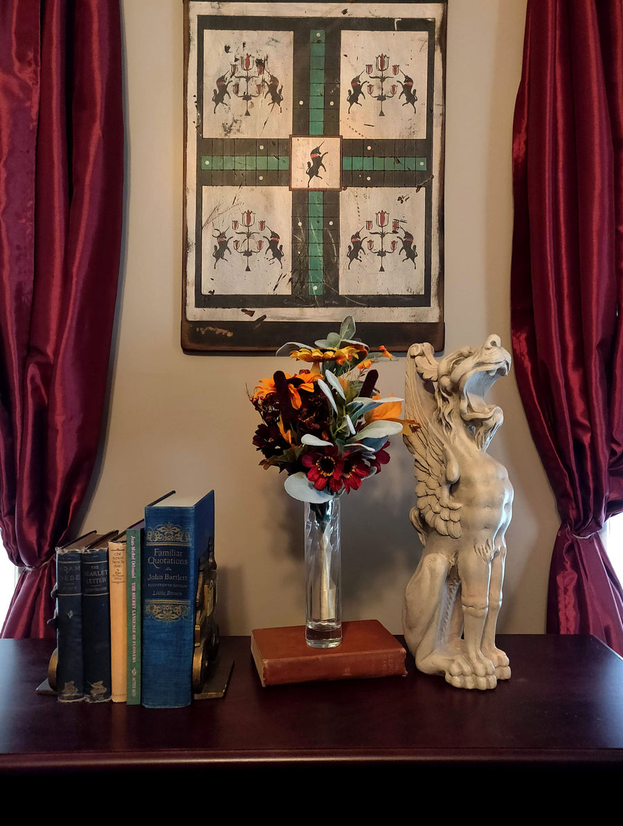 photo of plaster cast sculpture of ornamental seated winged lion on a dark sofa table with antique books and flowers, red curtains behind, and painting on wall