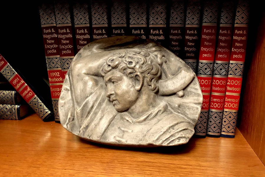 photo of plaster cast sculpture relief of upper body of male with arm raised behind head and holding a hammer leaning against an encyclopedia series in a wooden bookshelf