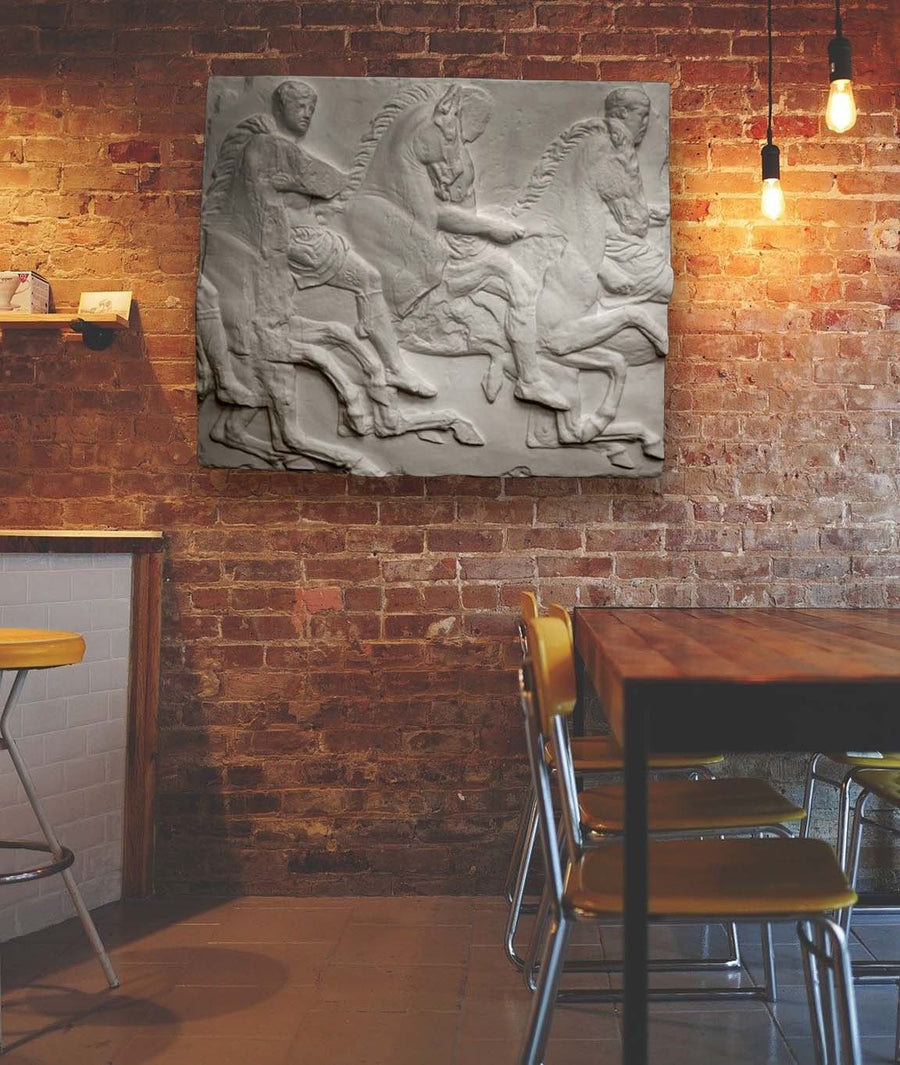 Photo of plaster cast relief sculpture of men on horses from Parthenon Frieze on brick wall with table and chairs on right and bar on left, cropped