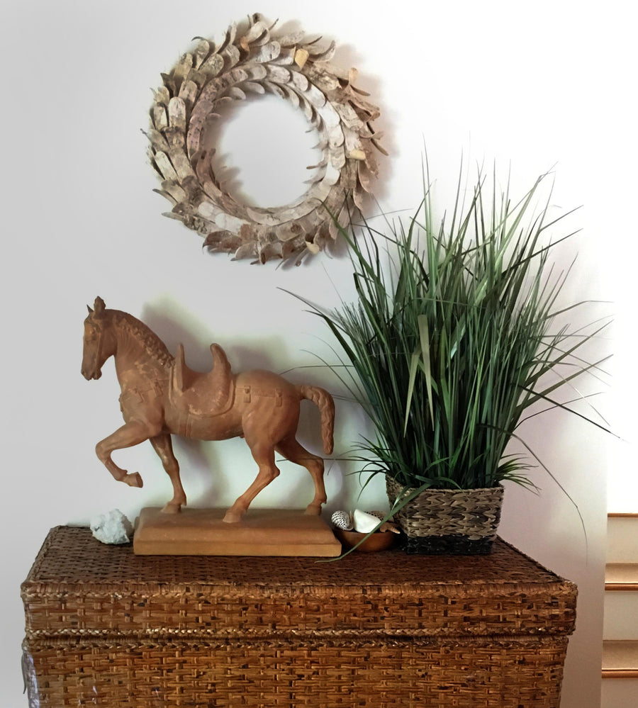 Photo of a plaster cast of a horse with a terra cotta finish on a wicker cabinet with a plant to the right and a metal wreath on the wall above