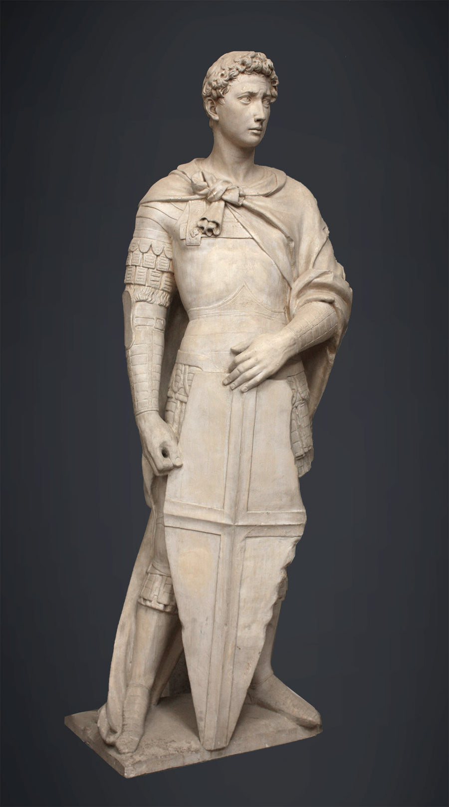 photo of plaster cast sculpture of male figure, namely Saint George, standing in armor and resting shield in front, on dark gray background