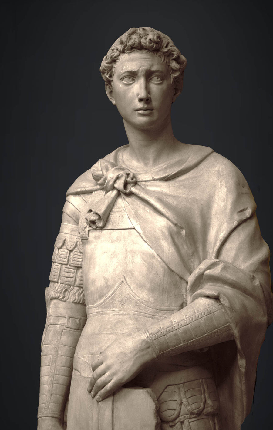 photo detail of plaster cast sculpture of male figure, namely Saint George, standing in armor and resting shield in front, on dark gray background