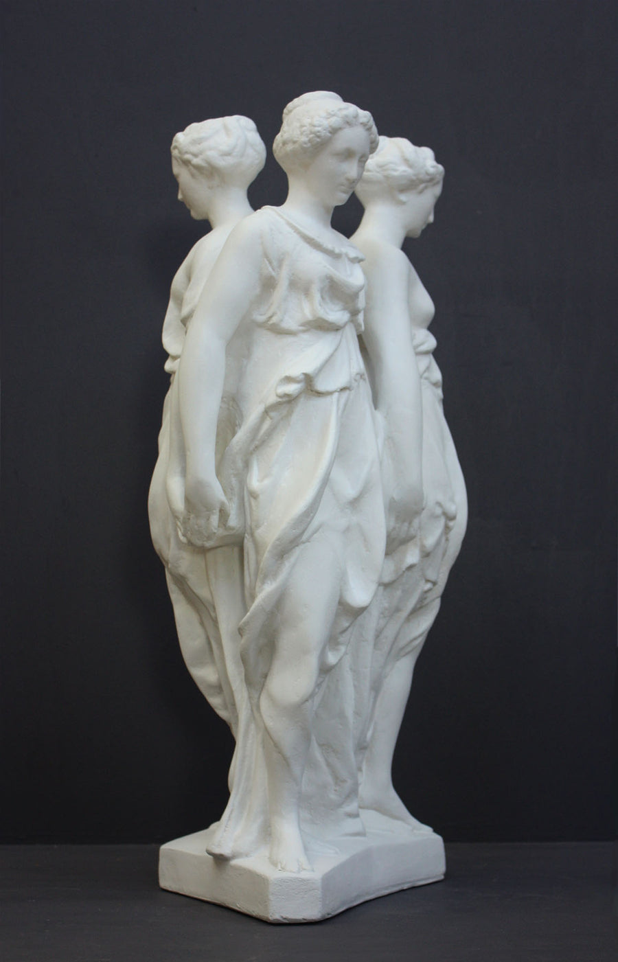 photo of white plaster cast sculpture of three robed females standing in a circle with their backs to each other against a gray background