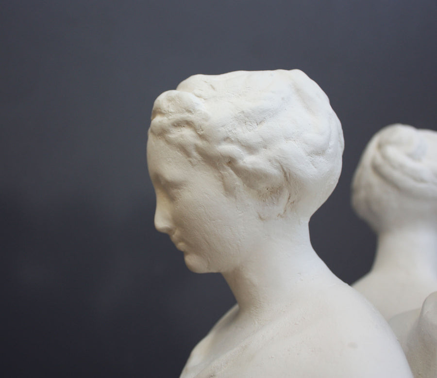 closeup photo of female head in profile from white plaster cast sculpture of three robed females standing in a circle with their backs to each other against a gray background