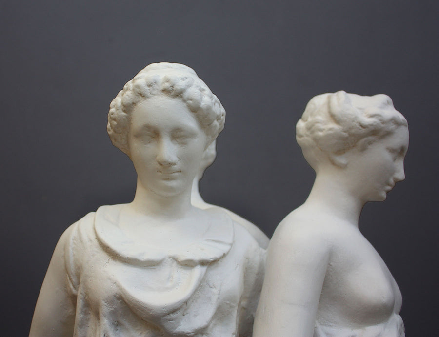 closeup photo of two female heads from white plaster cast sculpture of three robed females standing in a circle with their backs to each other against a gray background