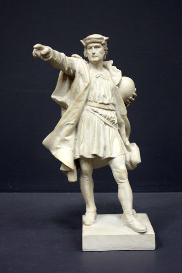 photo of plaster cast sculpture of standing man, namely Christopher Columbus, in robes and hat, pointing with right hand and holding a globe in his left arm against gray background