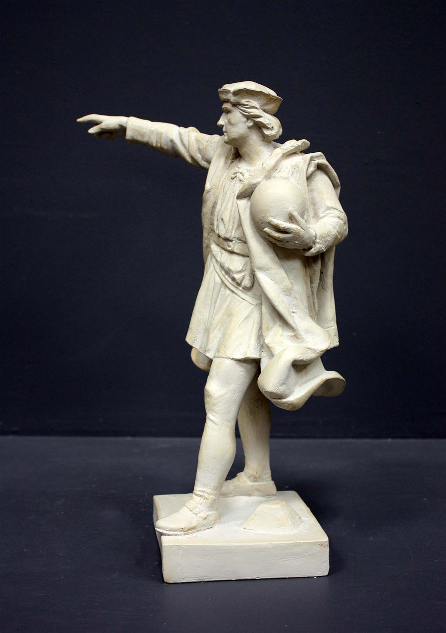 photo of plaster cast sculpture of standing man, namely Christopher Columbus, in robes and hat, pointing with right hand and holding a globe in his left arm against gray background