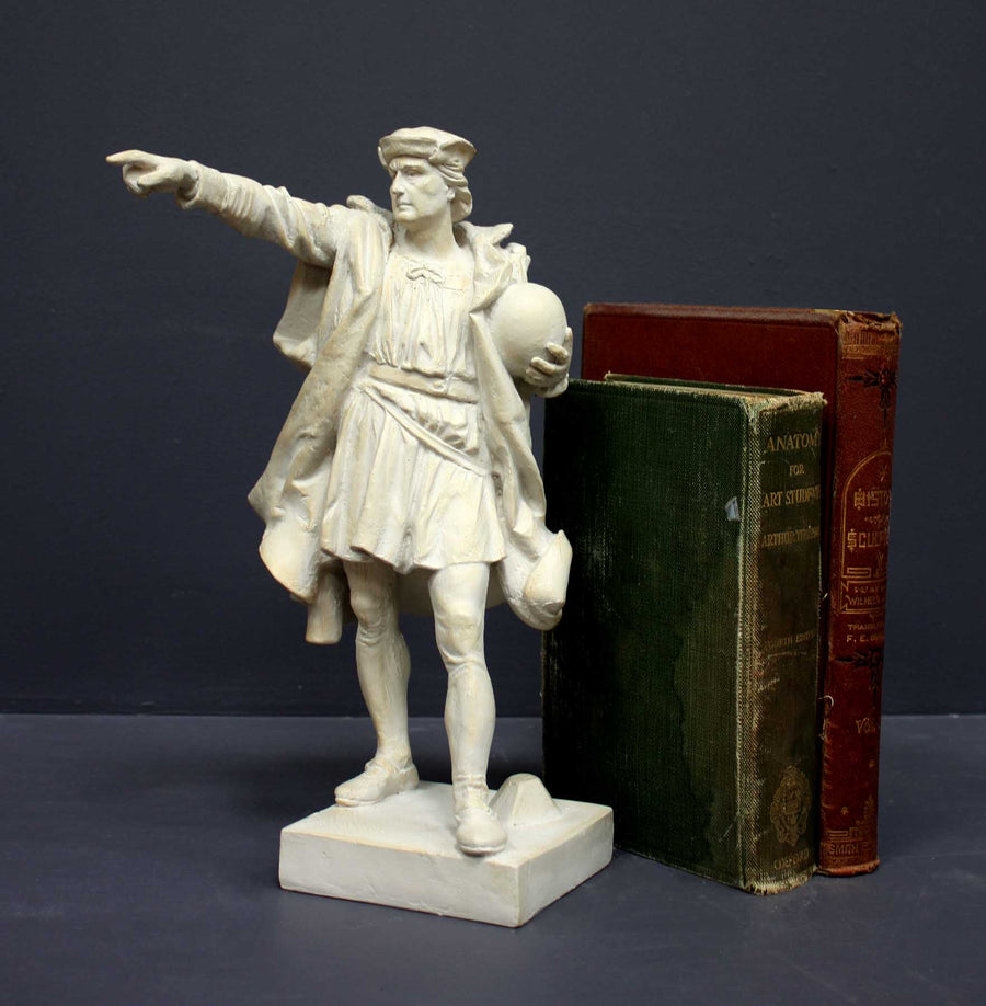 photo of plaster cast sculpture of standing man, namely Christopher Columbus, in robes and hat, pointing with right hand and holding a globe in his left arm with green and red books standing beside it against gray background