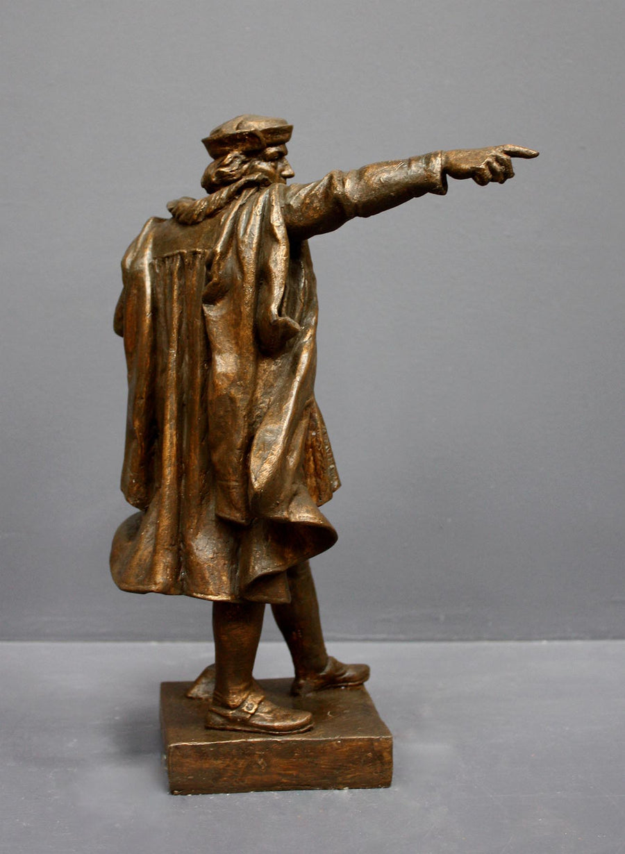 photo of bronze-colored plaster cast sculpture of standing man, namely Christopher Columbus, in robes and hat, pointing with right hand and holding a globe in his left arm against gray background