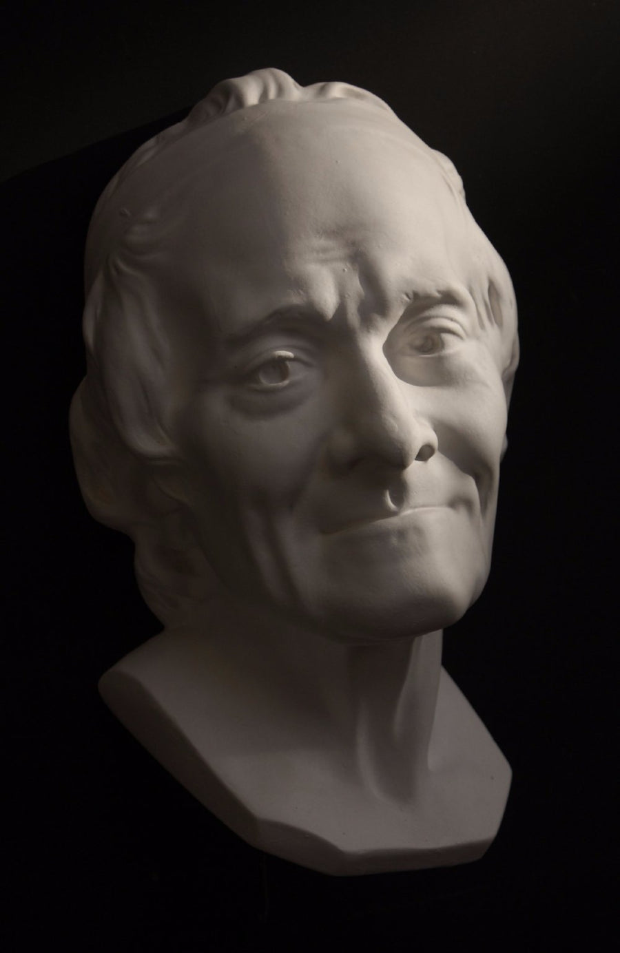 Photo with black background of plaster cast sculpture of elderly male head, namely Voltaire