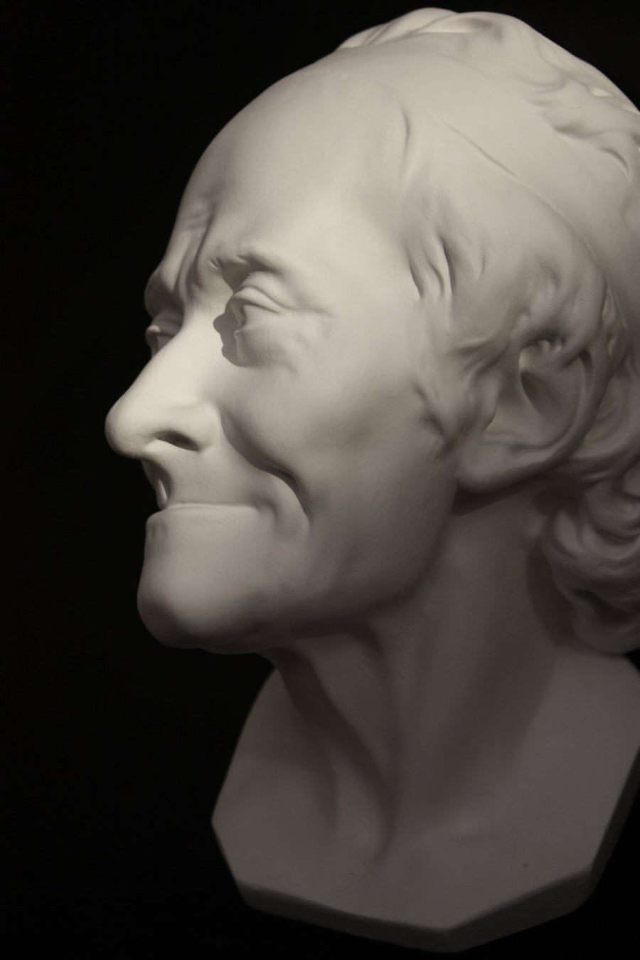 Photo with black background of plaster cast sculpture of elderly male head, namely Voltaire