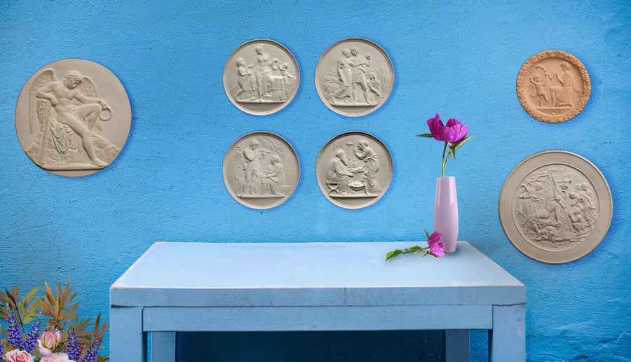 close up of light blue desk with small pink vase with pink flowers against bright blue wall featuring seven off-white plaster cast relief sculptures