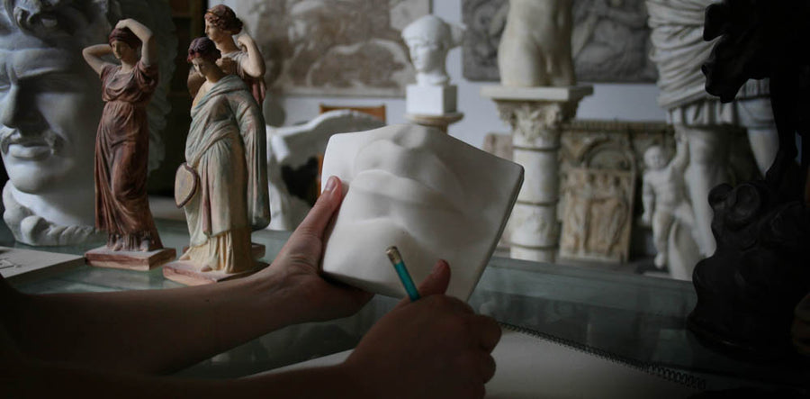 Photo of a woman's left hand holding the plaster cast of David's mouth by Michelangelo and right hand holding a drawing pencil with sculptures and a sketchpad in the background