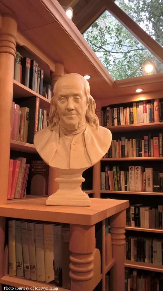 Photo of male bust sculpture on tall shelf with book shelves behind it